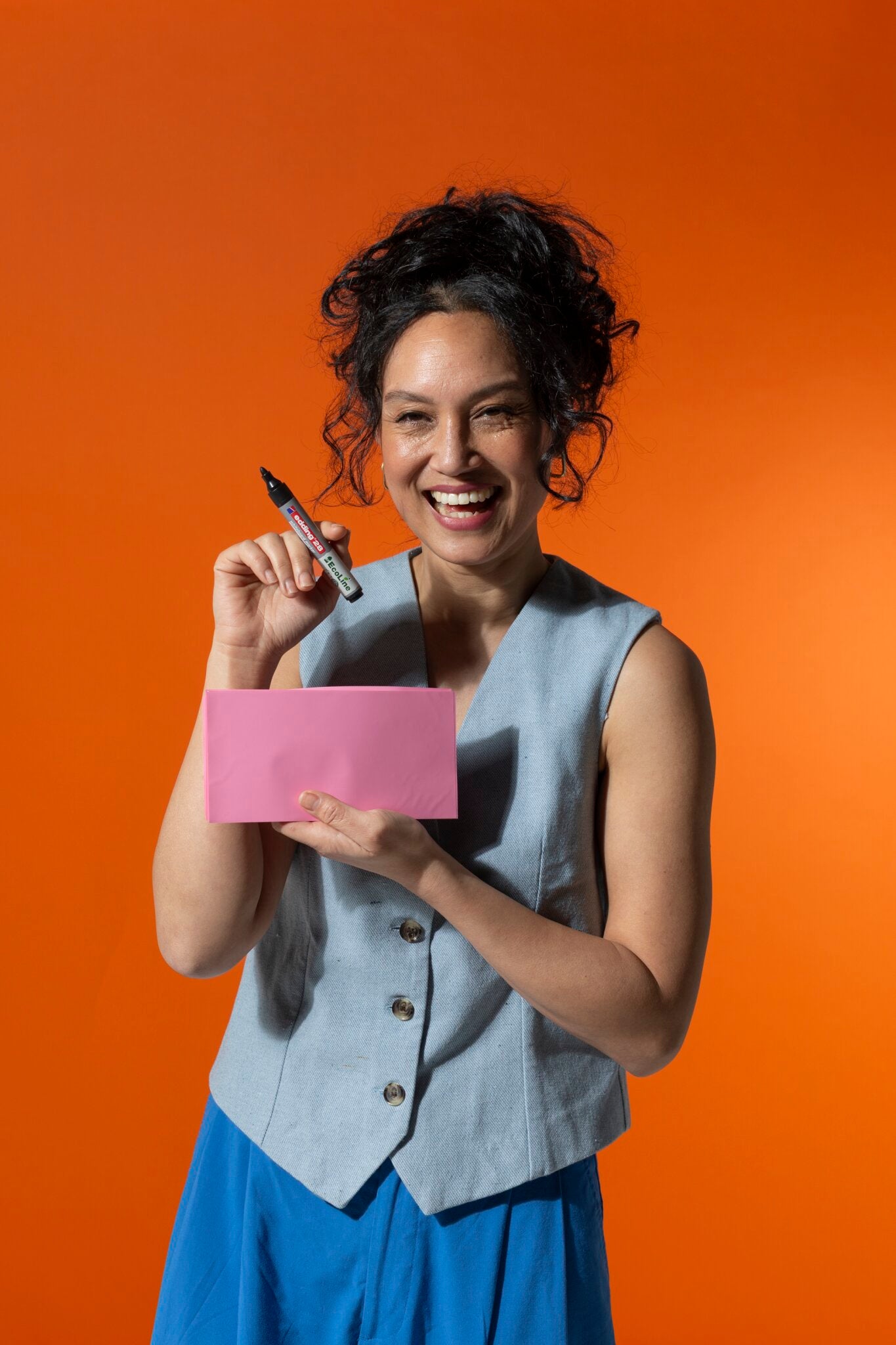 A smiling woman holds a PLAYROOM ESSENTIALS creative box Magic-Chart and a marker against an orange background, ready for organized brainstorming, dressed in a light blue vest and blue pants.