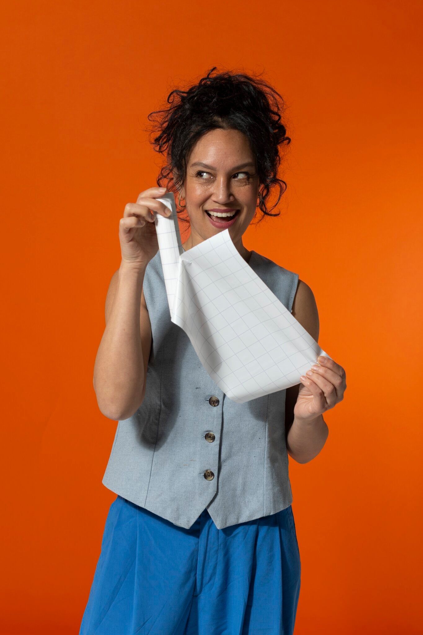 A smiling woman in a light gray vest and blue pants peels a sheet of white PLAYROOM ESSENTIALS creative box Magic-Chart by PLAYROOM against an orange background—ideal for organized brainstorming.