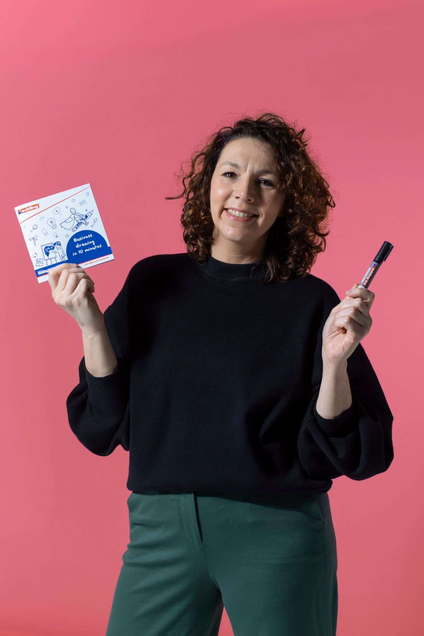 A woman with curly hair, dressed in a black top and green pants, smiles while holding a marker and PLAYROOM ESSENTIALS creative box Visualisation by PLAYROOM, highlighting her creativity against a pink background.