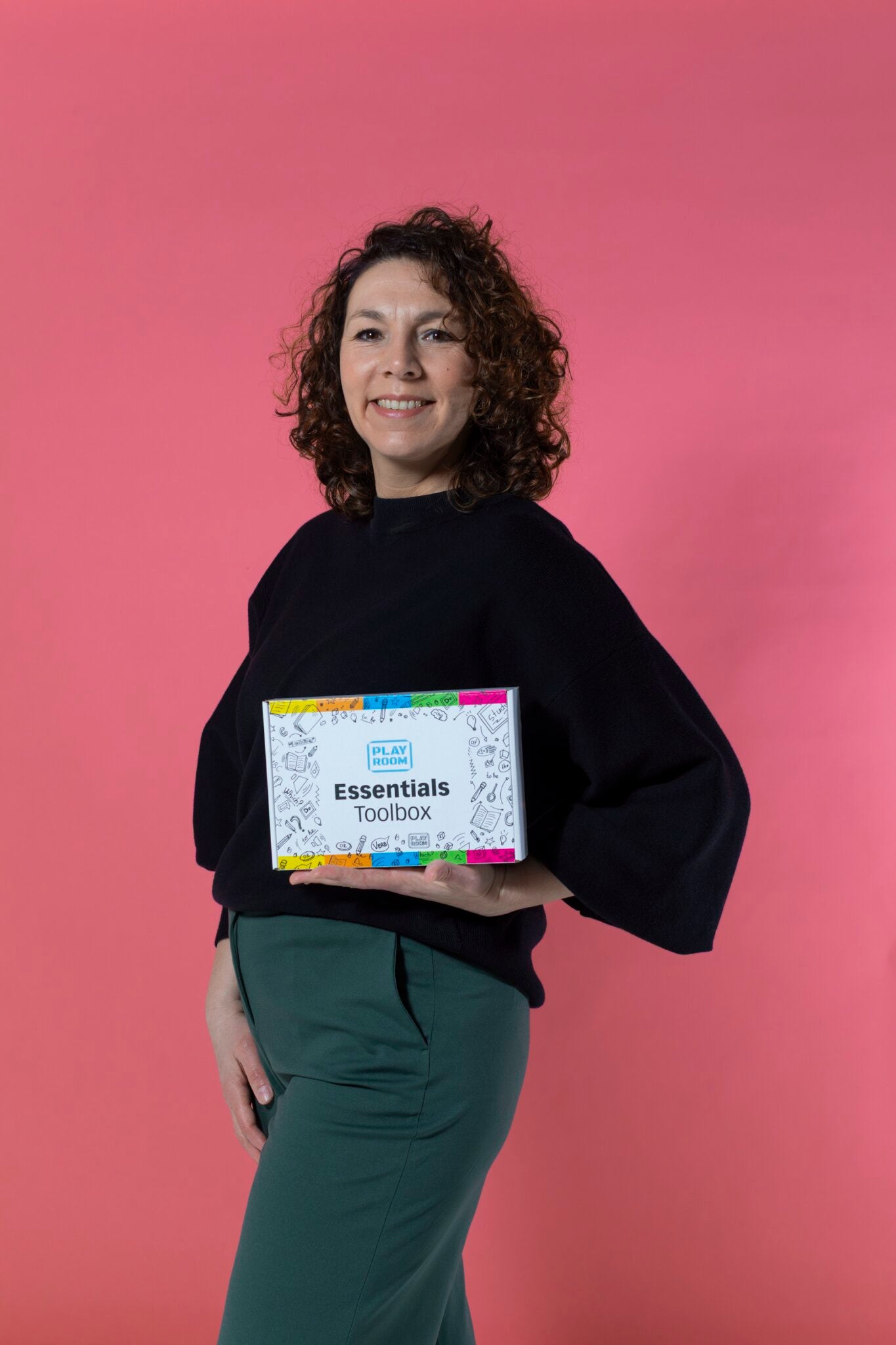 A woman with curly hair smiles against a pink background, holding PLAYROOM's "Essentials creative box Visualisation"—the perfect kit for enhancing creativity and visual communication.