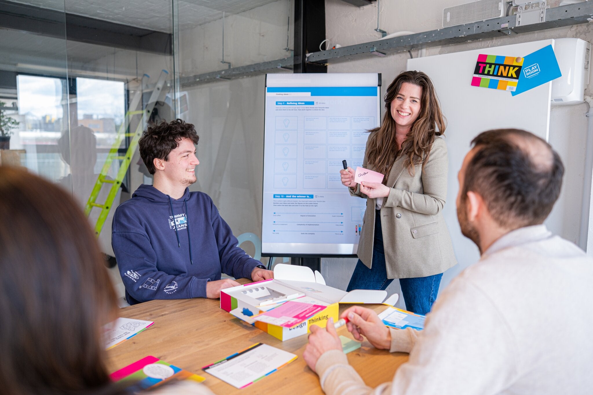 In a modern office, three people sit at a table while a woman leads a discussion by the whiteboard, using Legamaster Shop's PLAYROOM THINK FLIP starter set. Colorful materials and a "THINK" sign add to the creative atmosphere.