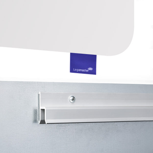 Close-up of a whiteboard mounted on a wall with a white metal rail and screw. A blue label with the Legamaster logo is partially visible behind the whiteboard.