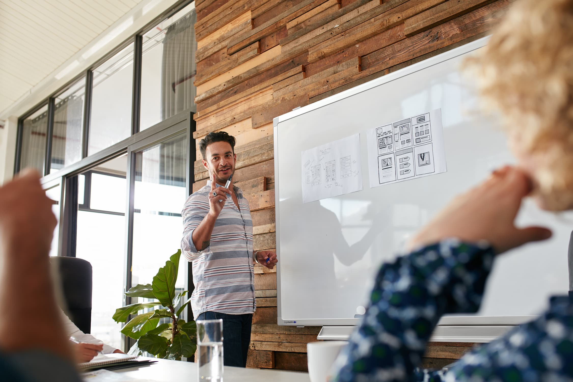 Three people in business attire stand by a whiteboard with sticky notes, having a discussion in a modern office with large windows and natural light.