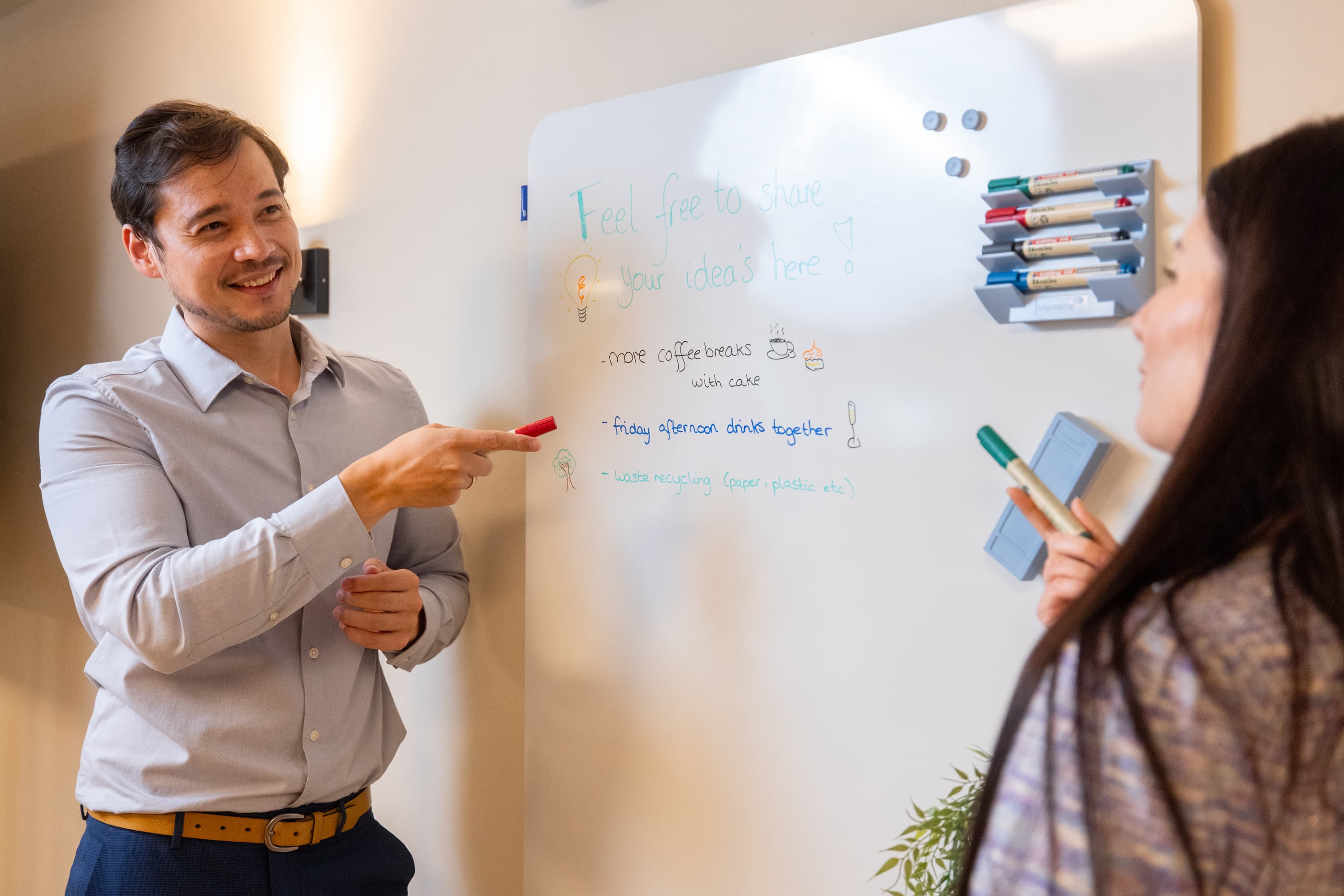 A man and a woman discuss ideas in front of a whiteboard with written notes and drawings. The man is smiling and pointing, while the woman holds a marker and listens.