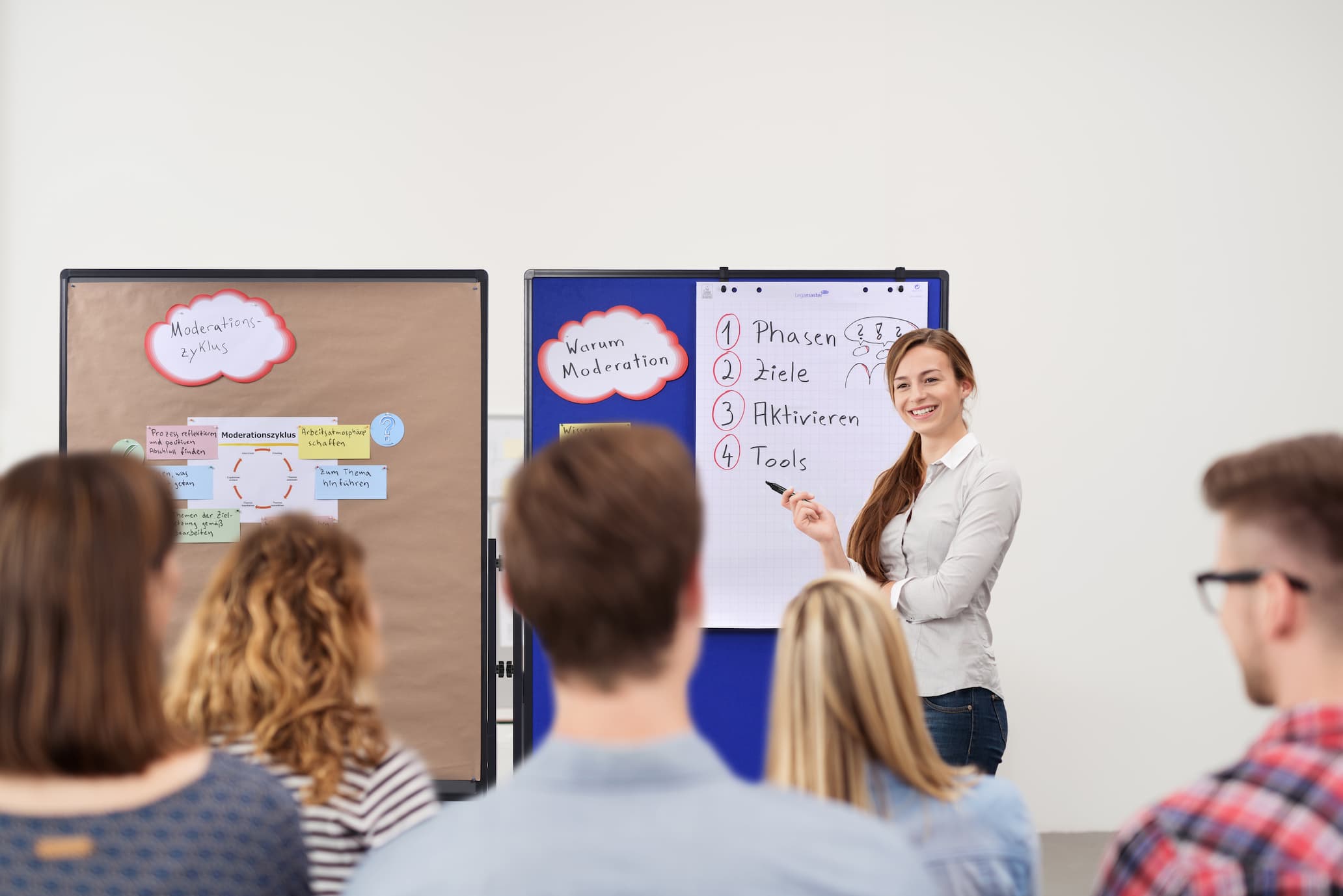 A woman stands by a flipchart presenting to a seated group. The flipchart lists four items in German, and a pinboard with diagrams is visible beside her. The setting appears to be a classroom or workshop.
