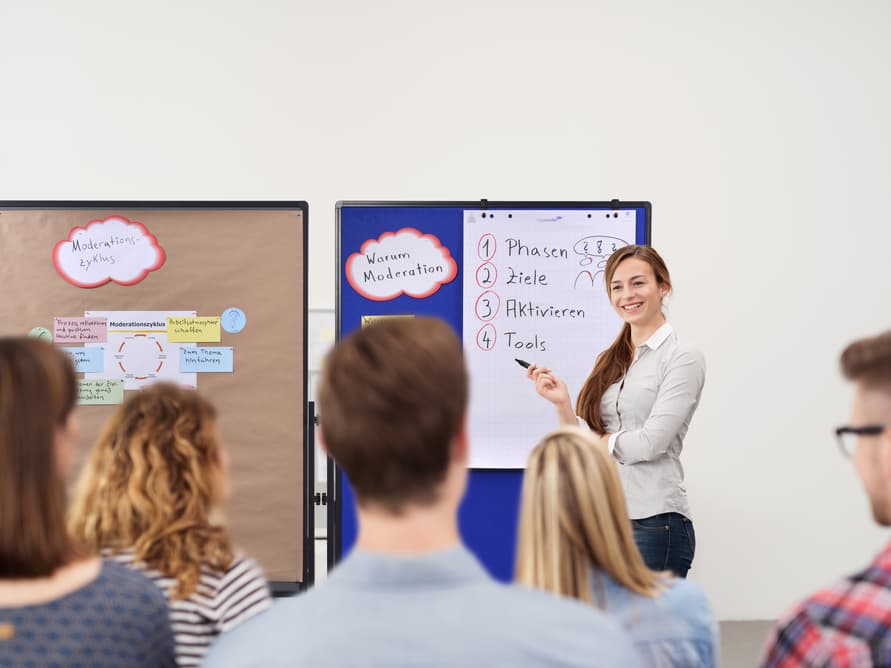 A woman stands by a flip chart, smiling and presenting to a seated audience. The flip chart has a numbered list in German. Another board with diagrams is visible beside her.