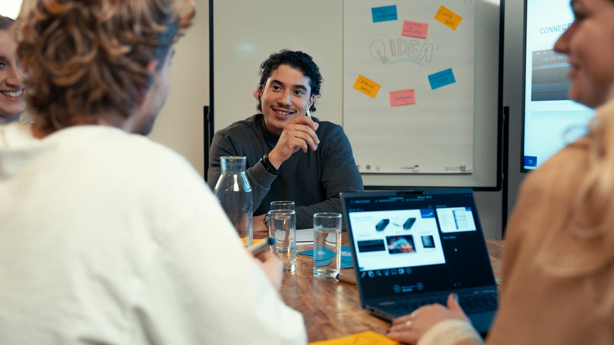 Three people sit around a table with laptops and water glasses, having a discussion. One person smiles while speaking. A whiteboard with colorful sticky notes is visible in the background.