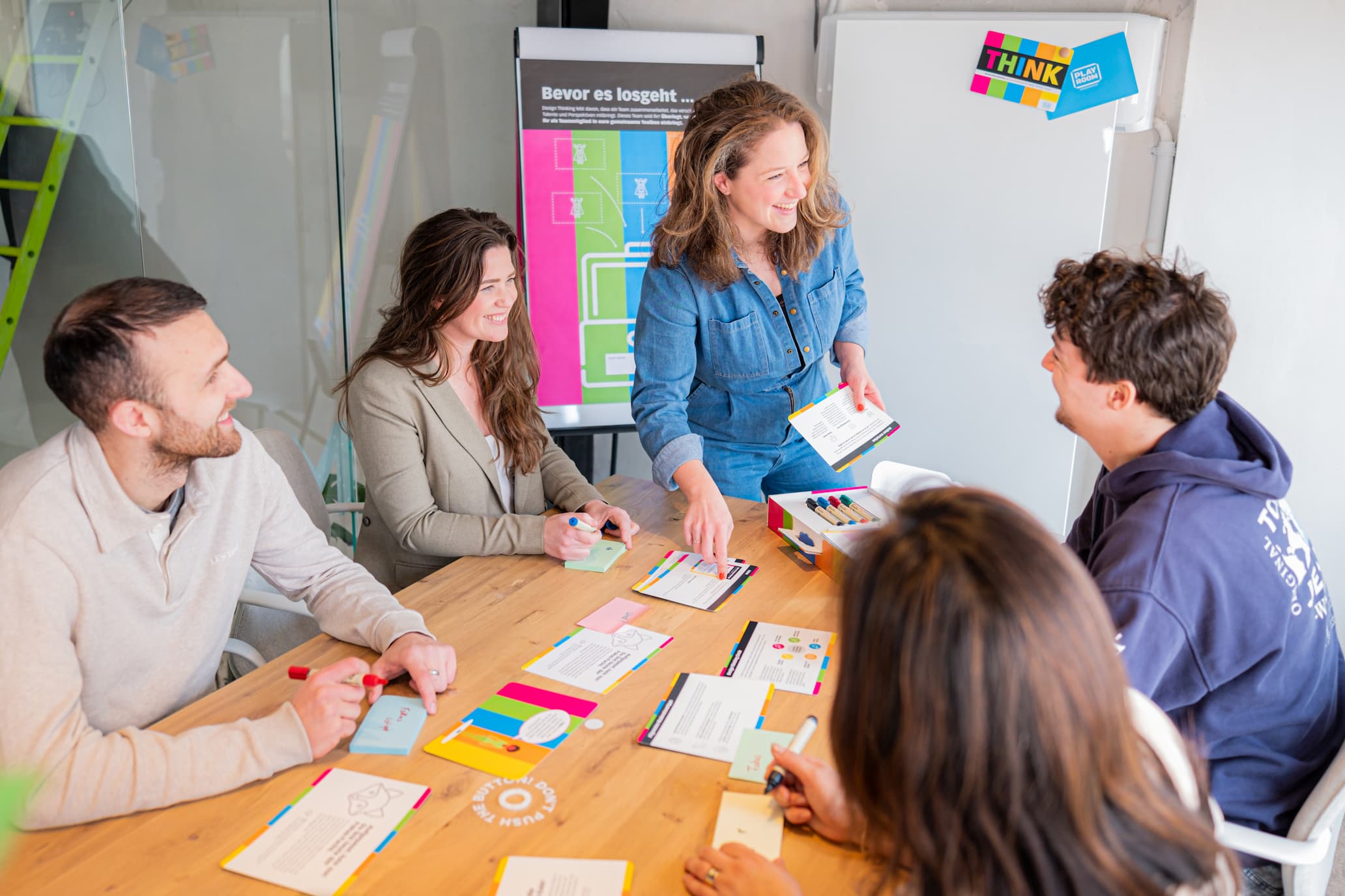 Four people sit around a table with colorful papers and markers, smiling and talking, while one woman stands and gestures, appearing to lead a creative workshop or brainstorming session.