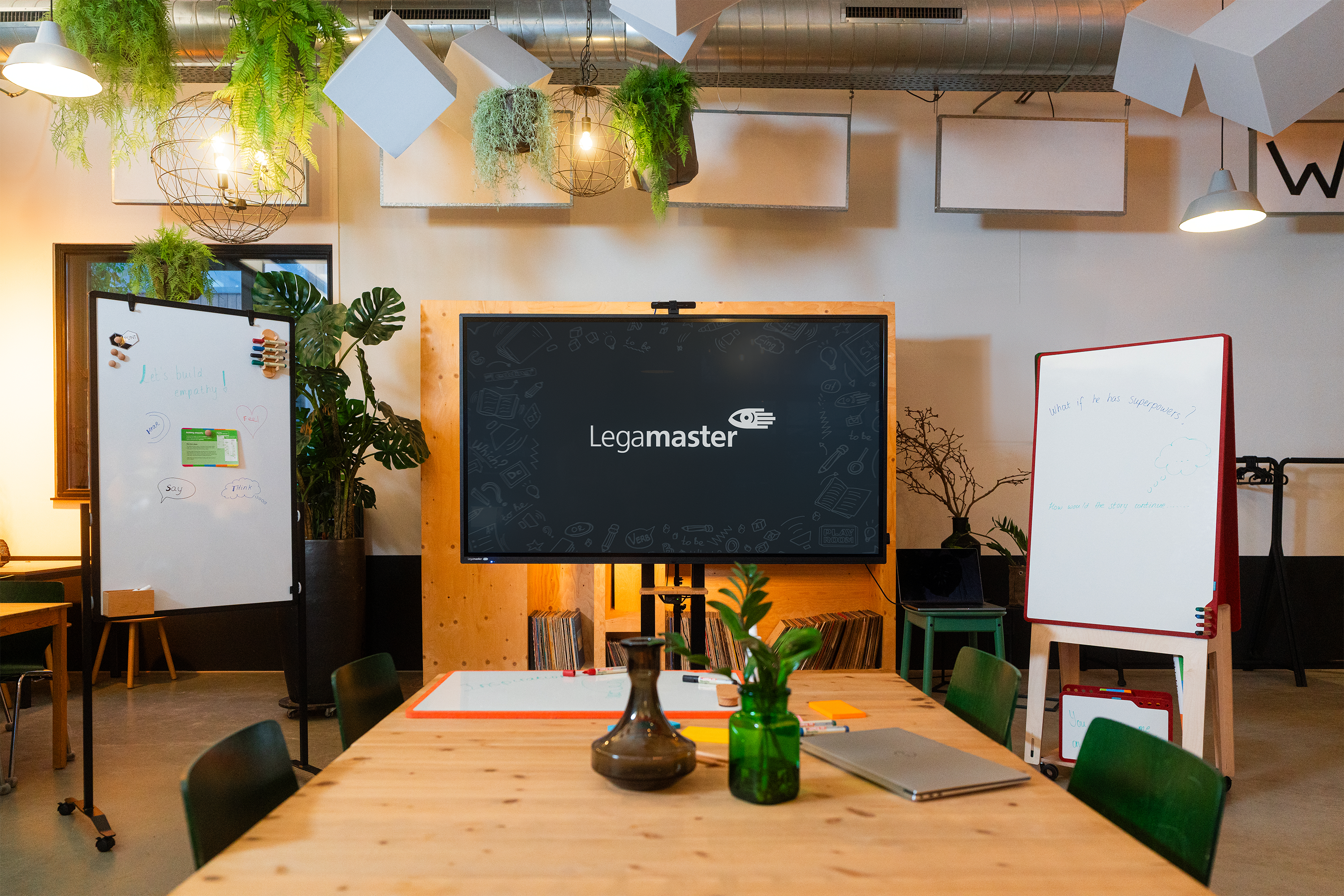 A modern meeting room with green chairs, a wooden table with vases, a large screen displaying "Legamaster," and two whiteboards with notes. Hanging plants and industrial-style lights decorate the space.