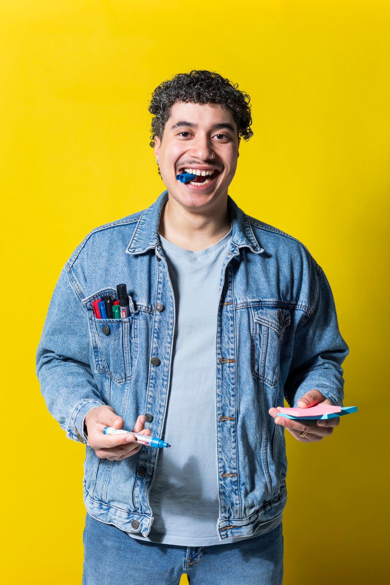 A smiling person with curly hair, wearing a denim jacket and light blue shirt, holds PLAYROOM ESSENTIALS creative box Workshop tools—markers and notepad—plus a blue marker in their mouth, set against a bright yellow background.
