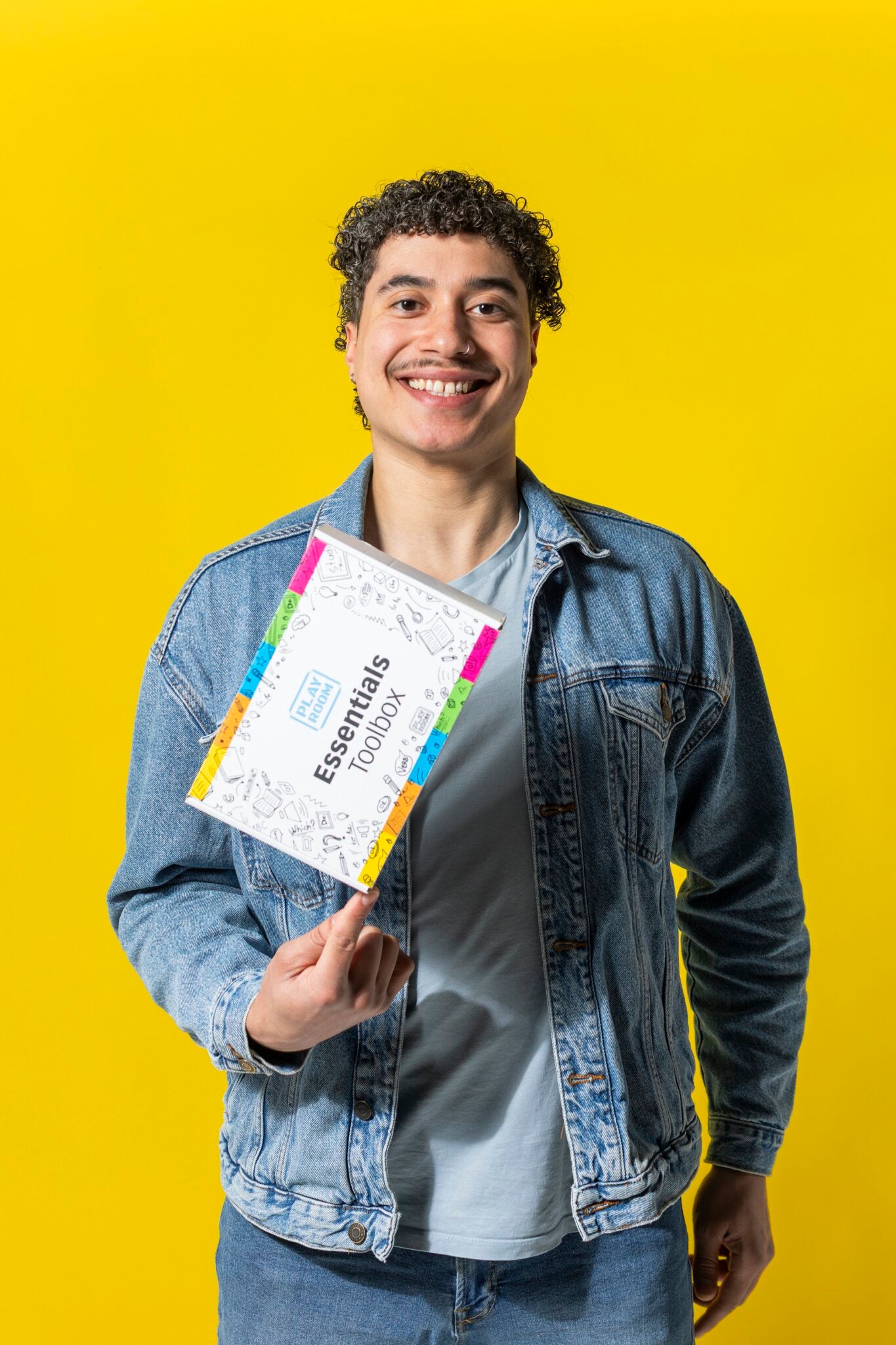 A smiling person in a denim jacket holds up the PLAYROOM ESSENTIALS creative box Workshop by PLAYROOM—a colorful productivity kit—against a bright yellow background.