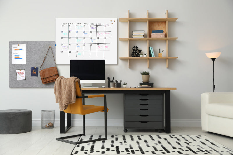A modern home office with a desk, computer, yellow chair, wall calendar, Legamaster BOARD-UP whiteboard for notes and bags, floating shelves with decor, a lamp, and a dotted rug on the floor.